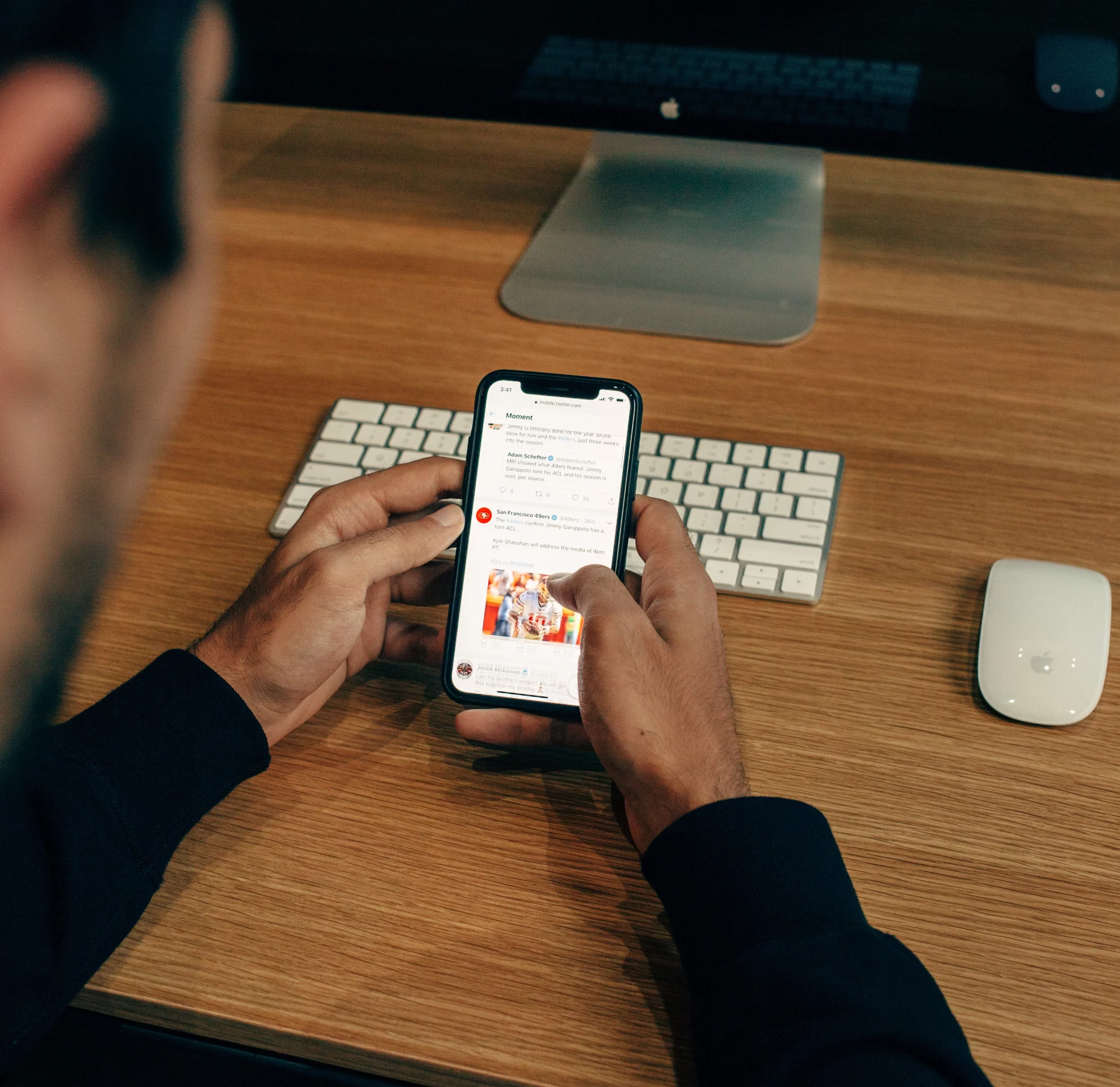 Das Bild zeigt eine Person, die ein Smartphone in den Händen hält und darauf eine Social-Media-App bedient. Der Mann, nur teilweise sichtbar mit einem Fokus auf seine Hände und das Smartphone, sitzt an einem modernen Arbeitsplatz mit einer hölzernen Tischplatte, auf der eine Apple-Tastatur, ein MacBook, und eine Apple-Maus zu sehen sind. Die Szene wird in warmen Tönen beleuchtet, was eine angenehme und professionelle Arbeitsatmosphäre schafft. Der Bildschirm des Smartphones zeigt verschiedene Social-Media-Posts, darunter scheinbar eine Nachricht und ein Foto von Kindern, was auf eine Interaktion mit sozialen Inhalten hinweist. Dieses Bild fängt einen alltäglichen Moment der digitalen Interaktion ein und stellt die Verbindung zwischen Technologie und alltäglicher Kommunikation dar.